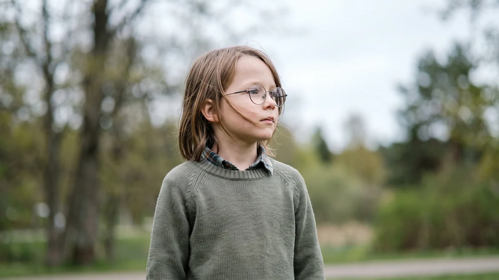 Et barn i skolealderen med briller står ude i et lyst nordisk parklandskab og kigger nysgerrigt mod horisonten