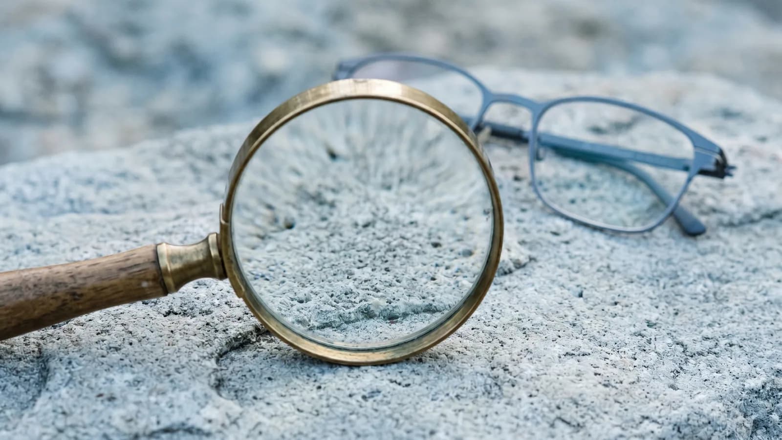 Et forstørrelsesglas der ligger på en kølig grå stenflade, med et par tynde briller ude af fokus i baggrunden