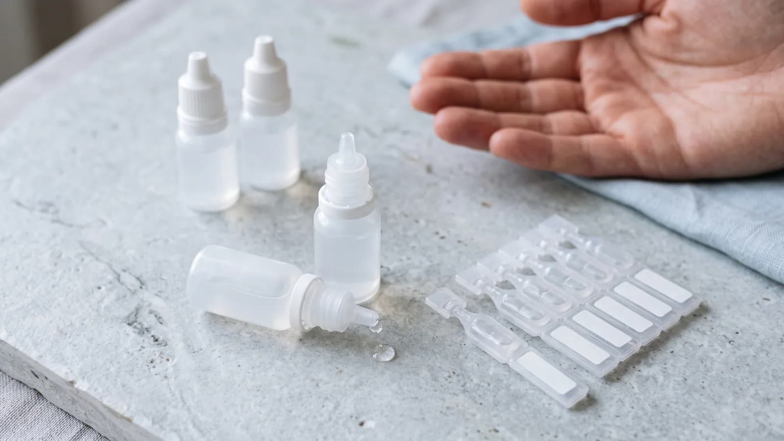 Flere umærkede øjendråbeflasker og en strip endosepipetter på en kølig lys stenflade, med en dråbe på spidsen