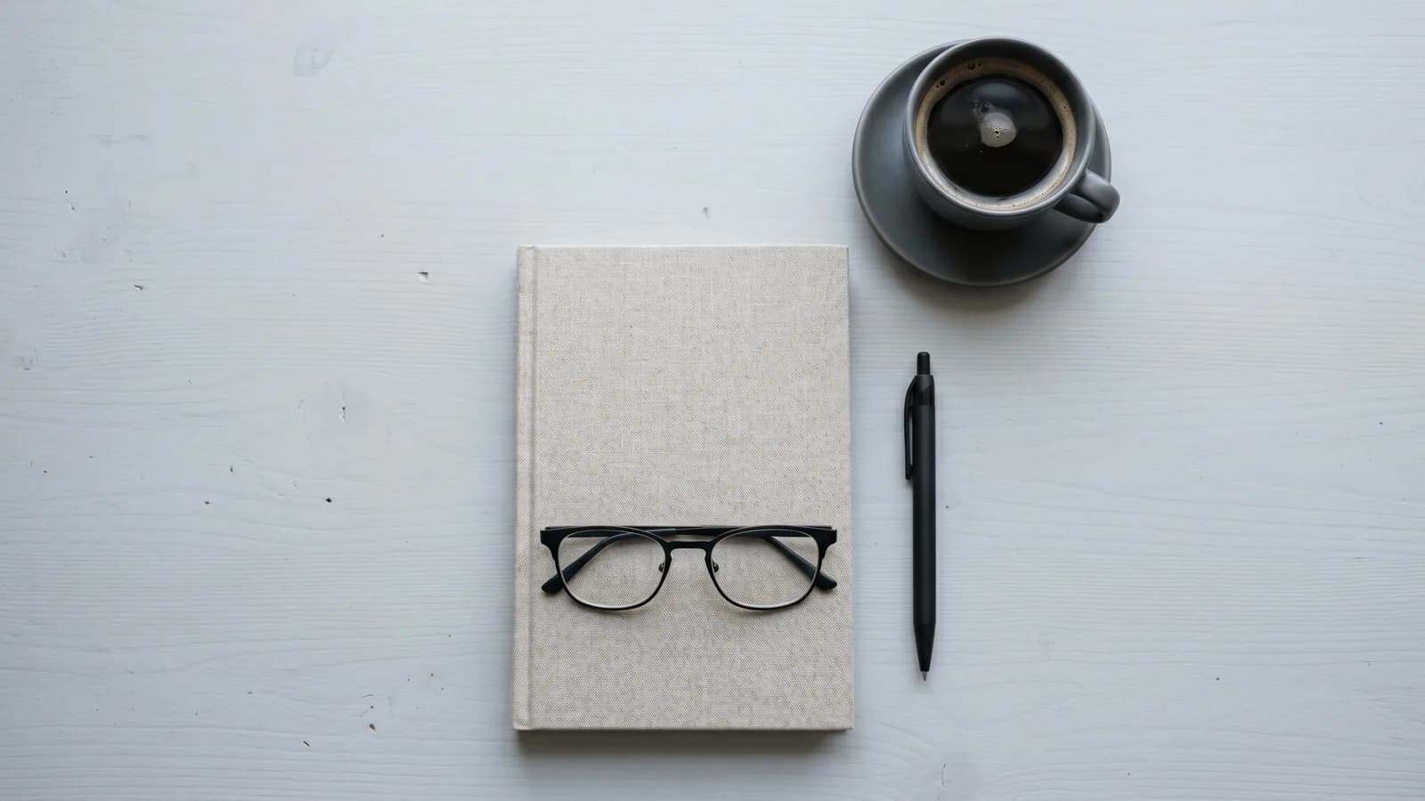 Briller på en lukket notesbog ved siden af en pen og en kop kaffe, set oppefra