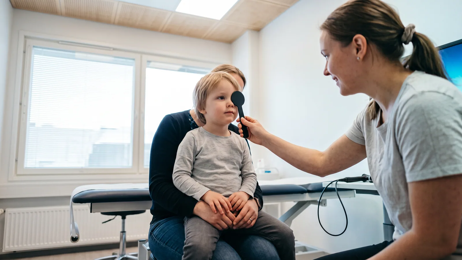 Et lille barn med briller kigger op, mens en øjenlæge lyser med en lille lommelygte mod barnets øjne i en rolig nordisk klinik