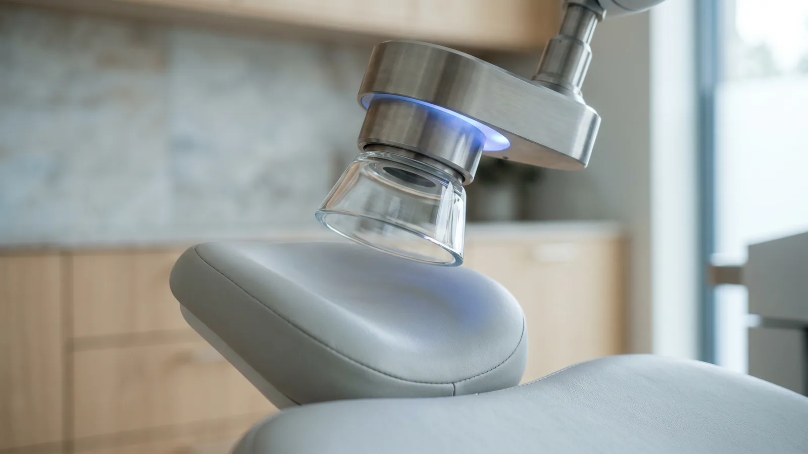 Nærbillede af dokningskeglen på en femtosekundlaser til øjenlaser, med et svagt blåt lys, over en tom patientstol i en lys nordisk øjenklinik.